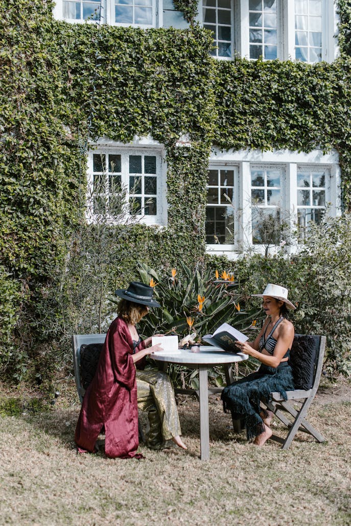 Two women enjoying a sunny day reading outdoors in a garden by an ivy-covered house.