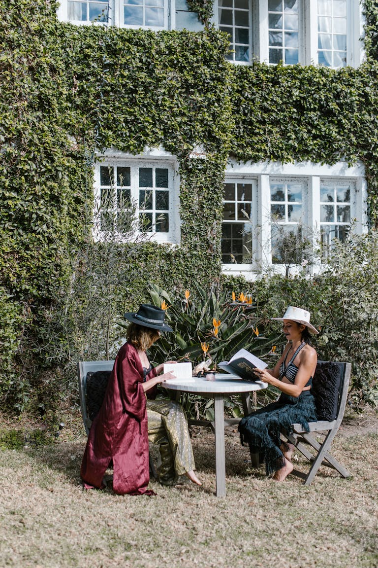 Two women enjoying a sunny day reading outdoors in a garden by an ivy-covered house.
