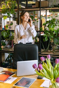 A woman smiling and talking on a phone in a bright, plant-filled office setting, standing by her desk.