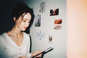 A young woman in a cozy room examining art prints on a wall with thoughtful expression.