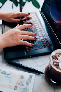 Close-up of a woman typing on a laptop in a cozy café with iced coffee nearby.