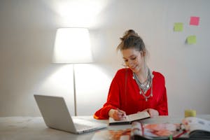 Smiling woman in red top writing in notebook at desk with laptop and lamp indoors.