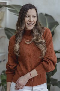 Young woman with wavy hair wearing a burnt orange sweater, smiling indoors with green plants.