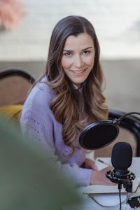 A smiling young woman recording a podcast at home with microphone and notebook.
