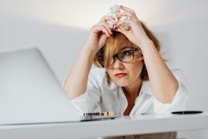 Frustrated woman with eyeglasses at a desk, crumpling paper while using a laptop indoors.