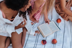 Teenage friends enjoying a sunny picnic with a book and peaches on a striped blanket.
