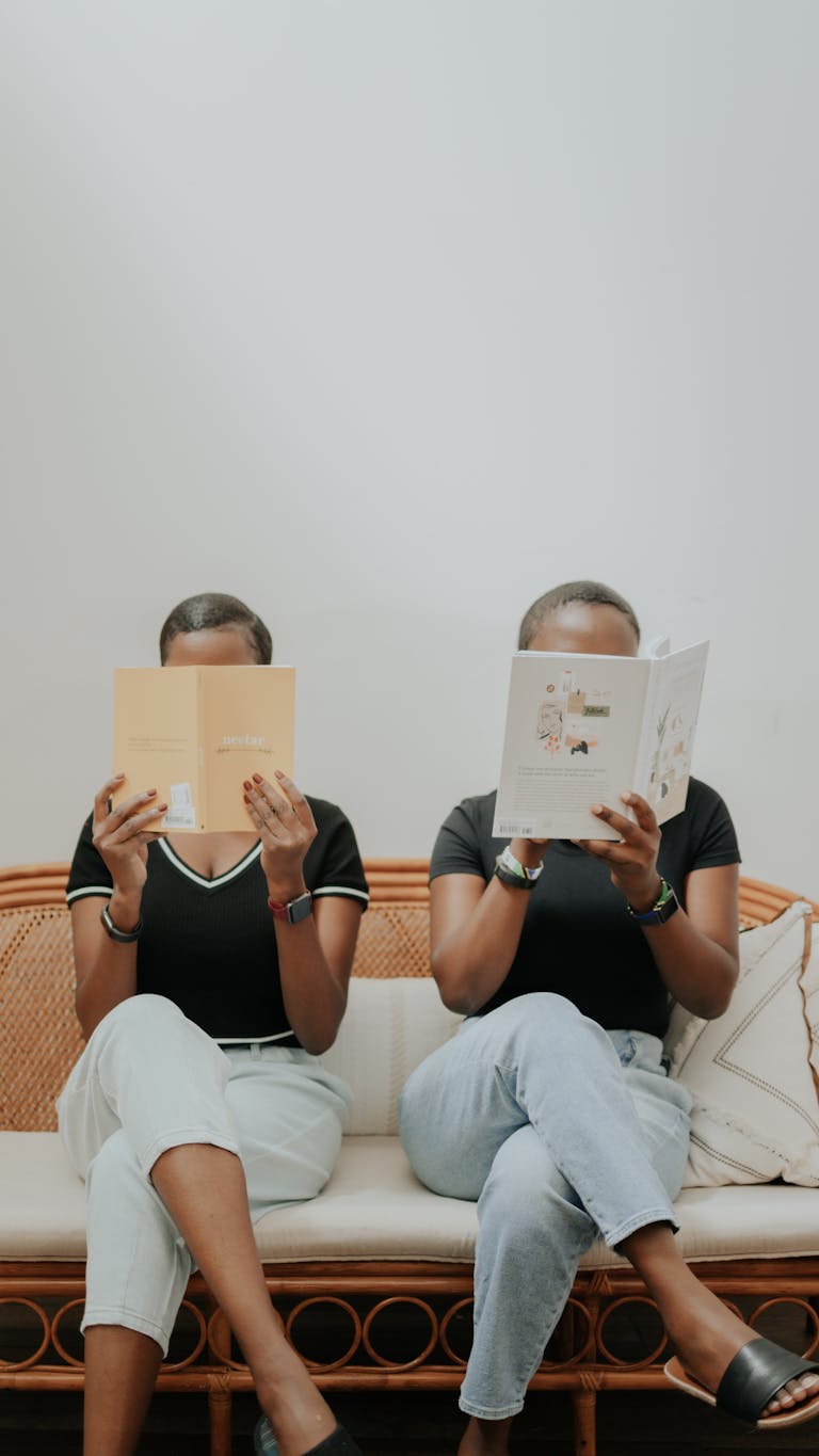 Two women sit on a rattan sofa reading books indoors, showcasing friendship and relaxation.