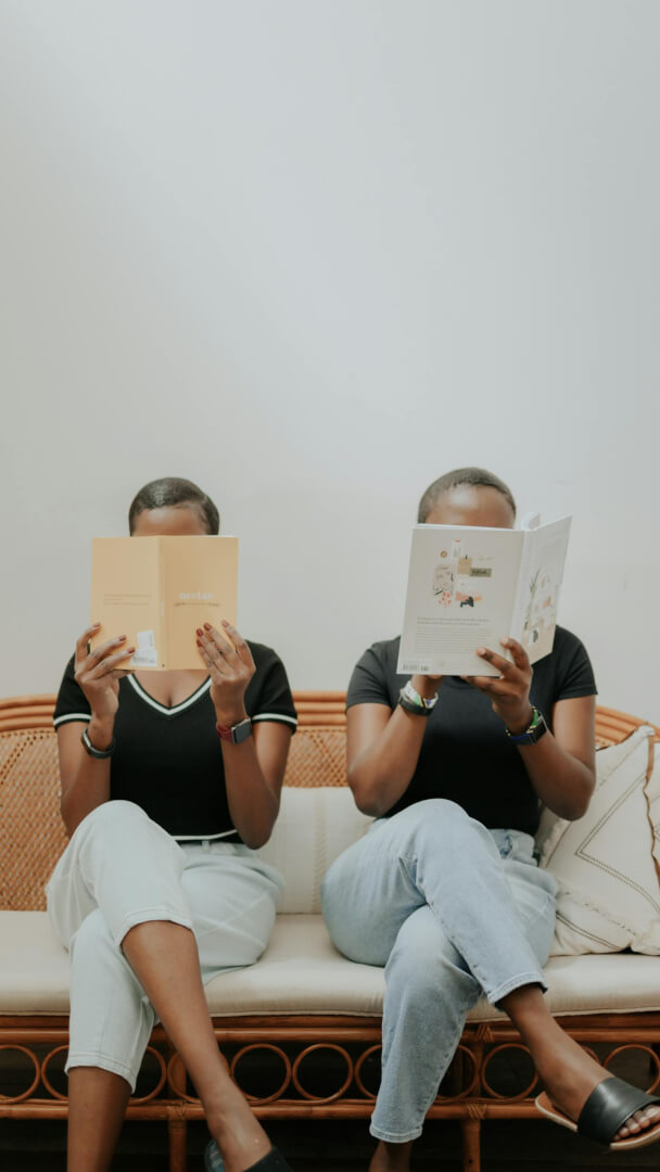 Two women sit on a rattan sofa reading books indoors, showcasing friendship and relaxation.